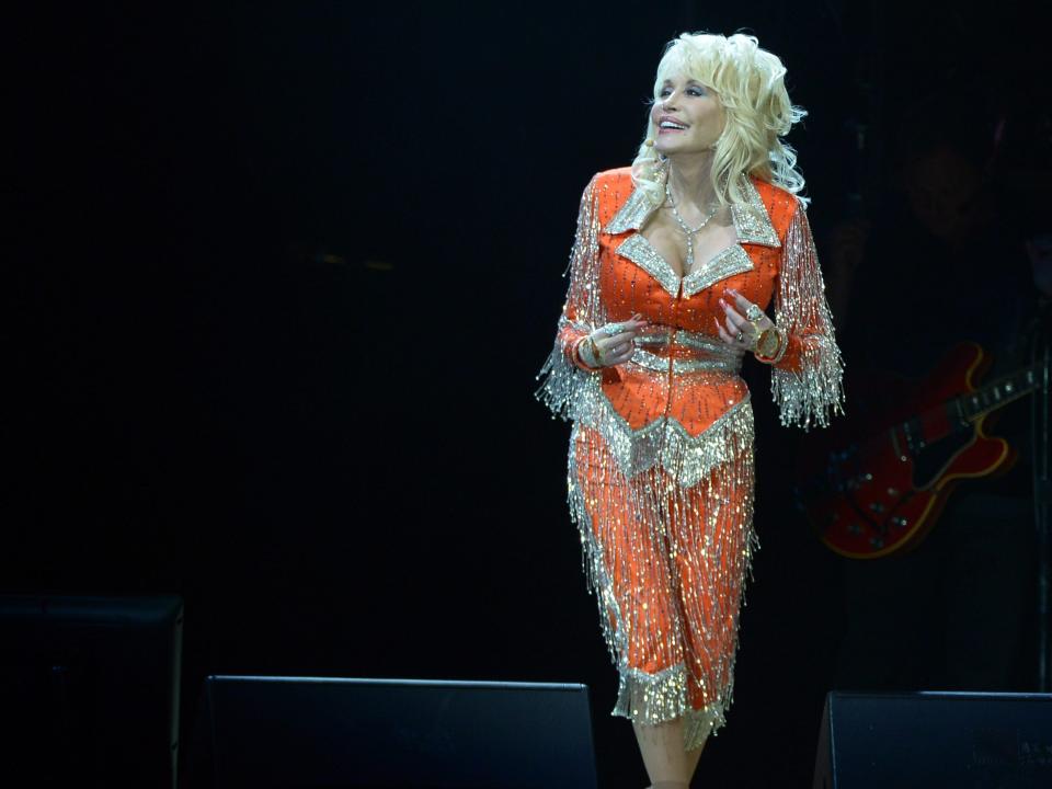 Dolly Parton on a dark stage wearing a bright orange and silver crystal embellished pantsuit costume.
