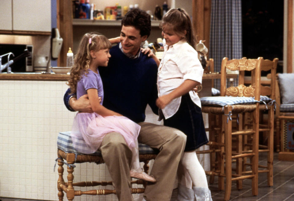 Jodie Sweetin sitting on Bob Saget's lap while Candace Cameron smiles.