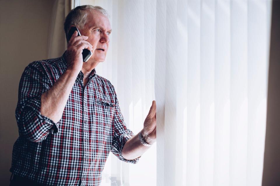 elderly man looking out window parting blinds and talking on cell phone