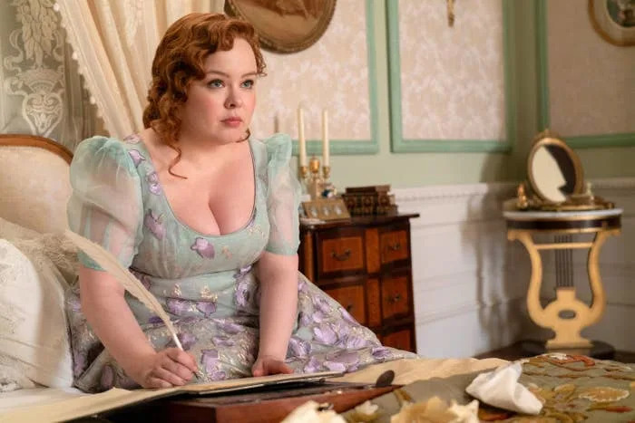 Nicola Coughlan dressed in a floral, puff-sleeved vintage gown, sits at a writing desk with a quill in hand in an elegant, historic interior setting