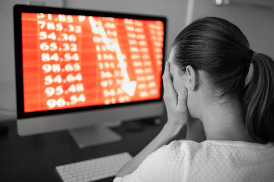 A person holding their face with a computer showing stock losses in the background.