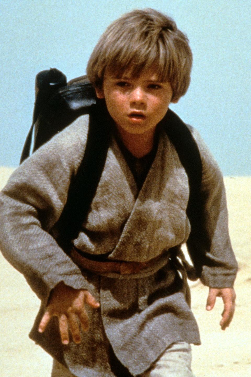 A young boy with short hair, wearing a robe and backpack, walks across a desert