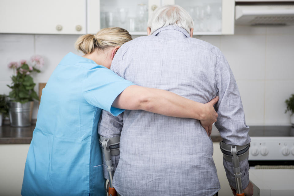 An aged care worker supports an elderly man on crutches.