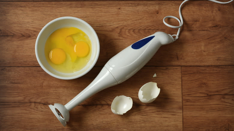 Immersion blender and eggs in bowl on wood surface
