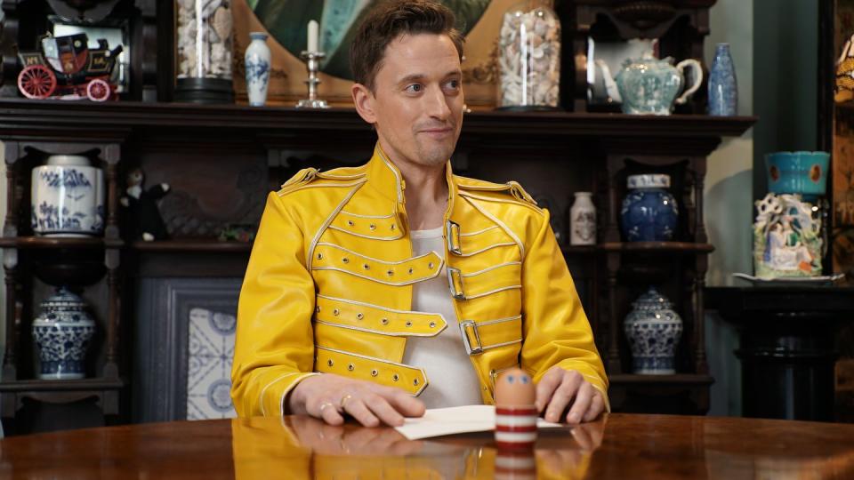 john robins sits inside the taskmaster house's task room, in front of a table containing a task and an egg in a striped cup with googly eye stickers on it