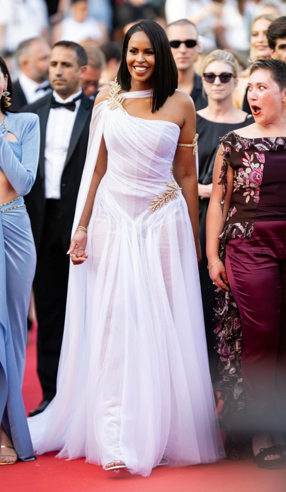 Sabrina in a one-shouldered white sheer gown with a flowing skirt and gold beaded palm leaves on her right shoulder and left hip.