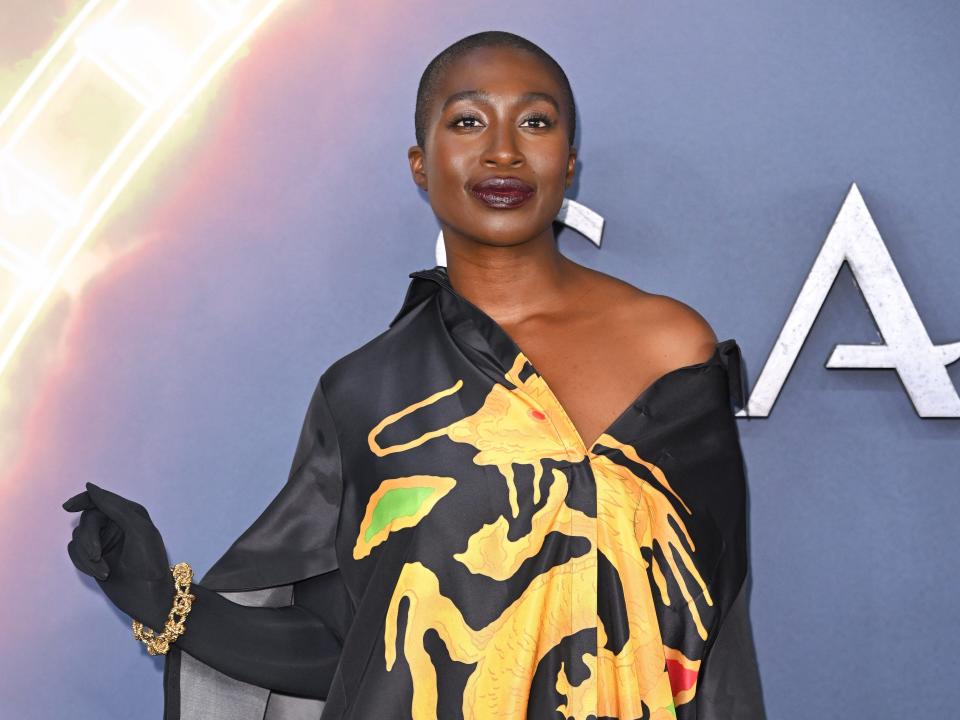 vivienne acheampong wearing a black off the shoulder long sleeve dress with a golden dragon printed on it. she's wearing long black gloves accented with gold bracelets, and is smiling slightly at the camera