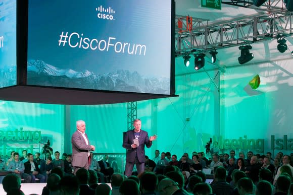 Two people in suits in front of an audience with a screen saying Cisco Forum above them.