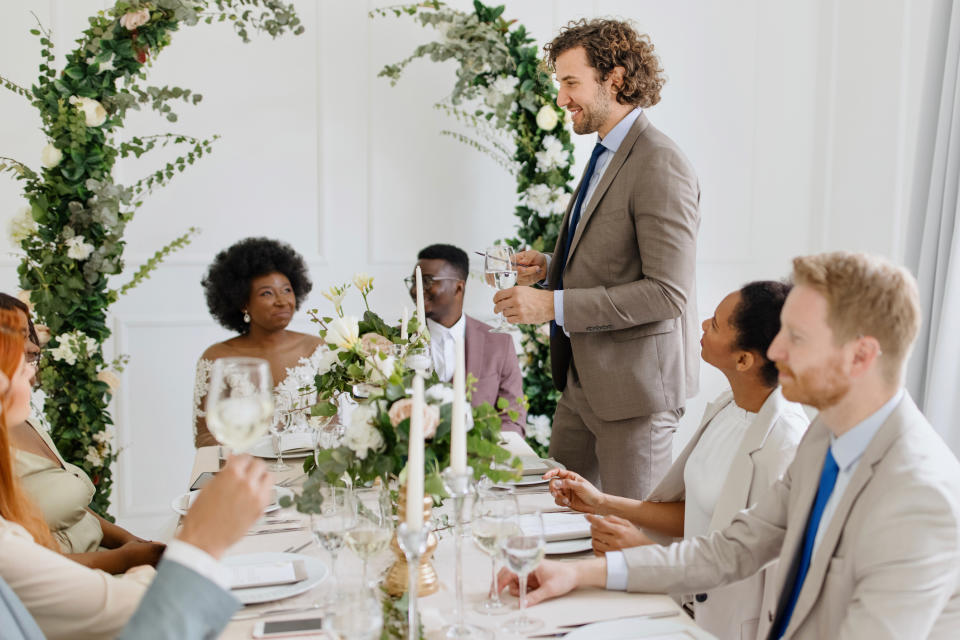 Guests at a wedding reception table listen to a man giving a toast