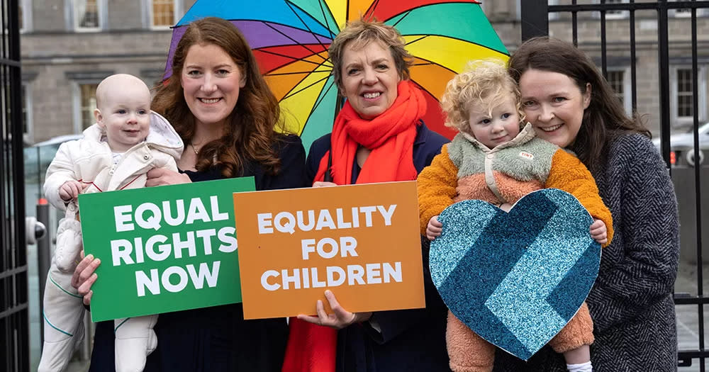 This article is about a new bill proposing amendments to ensure legal equality for children born to same-sex female parents. The image shows Ivana Bacik in the middle, with two women holding young children on either side of her. Ivana holds a rainbow umbrella above her head, and a sign reading 