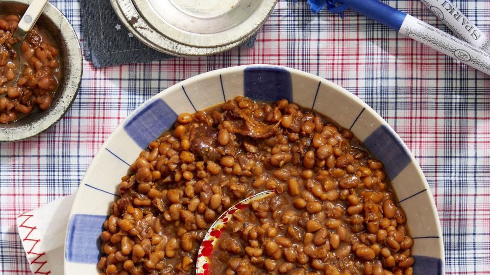 root beer baked beans in a serving bowl with a spoon on a wooden table with a red and blue plaid tablecloth