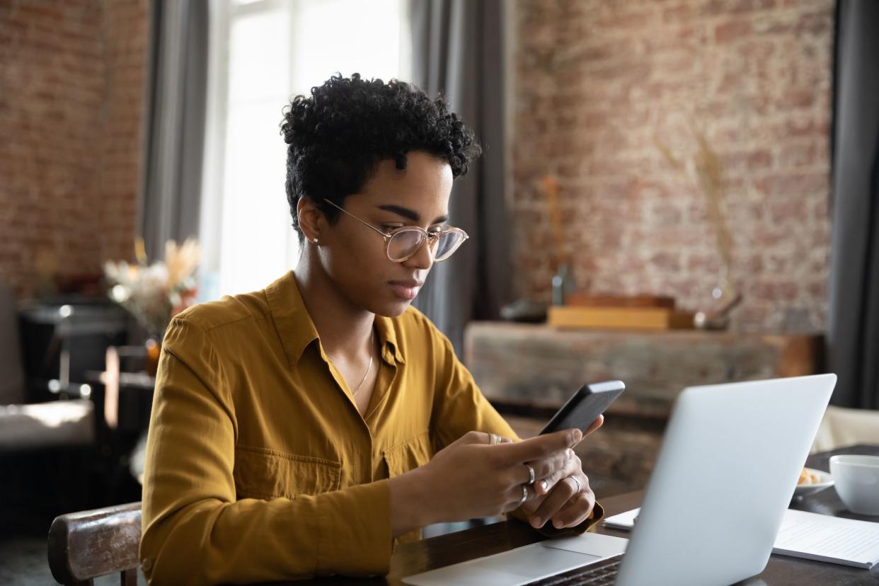 Young African businesswoman sit at workplace desk in front of laptop use smartphone, distracted from work on computer, using app chat online, synchronize files via application. Tech, workflow concept