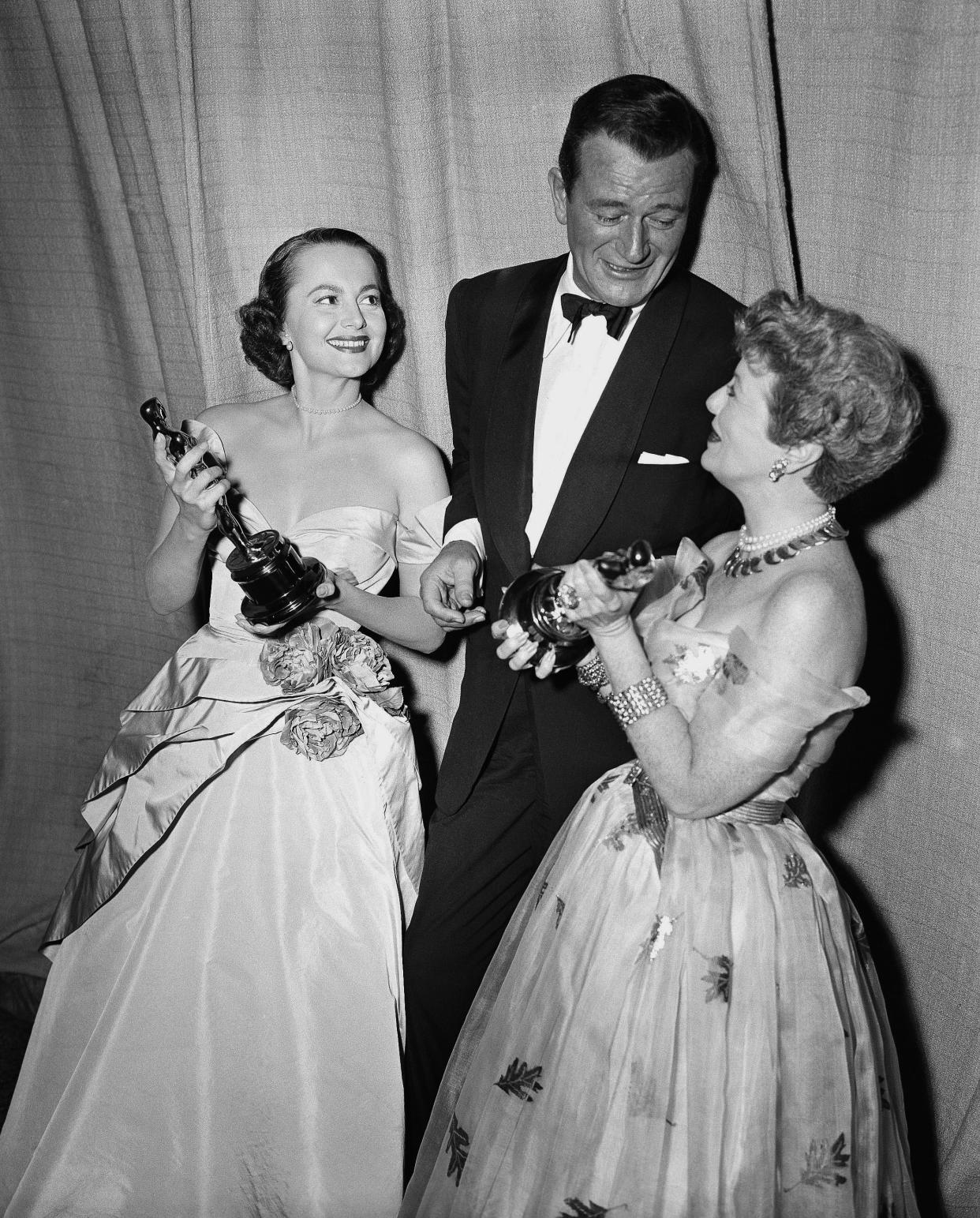 John Wayne shows the gold statuettes he accepted for Gary Cooper and John Ford to two former Academy Award winners, Olivia De Havilland and Janet Gaynor, after the ceremony in Hollywood on March 19, 1953.