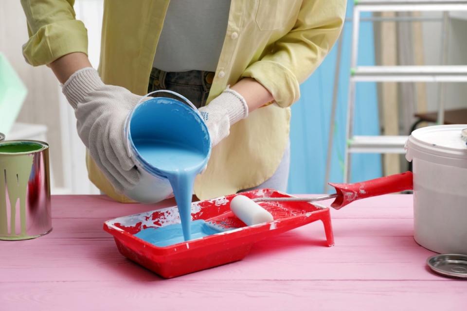 A DIY painter wearing cotton gloves pouring light-blue paint into a red paint tray.