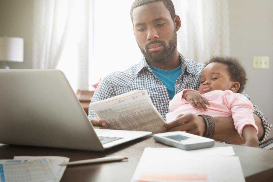 Father sitting at laptop, reading utility bill while holding baby.