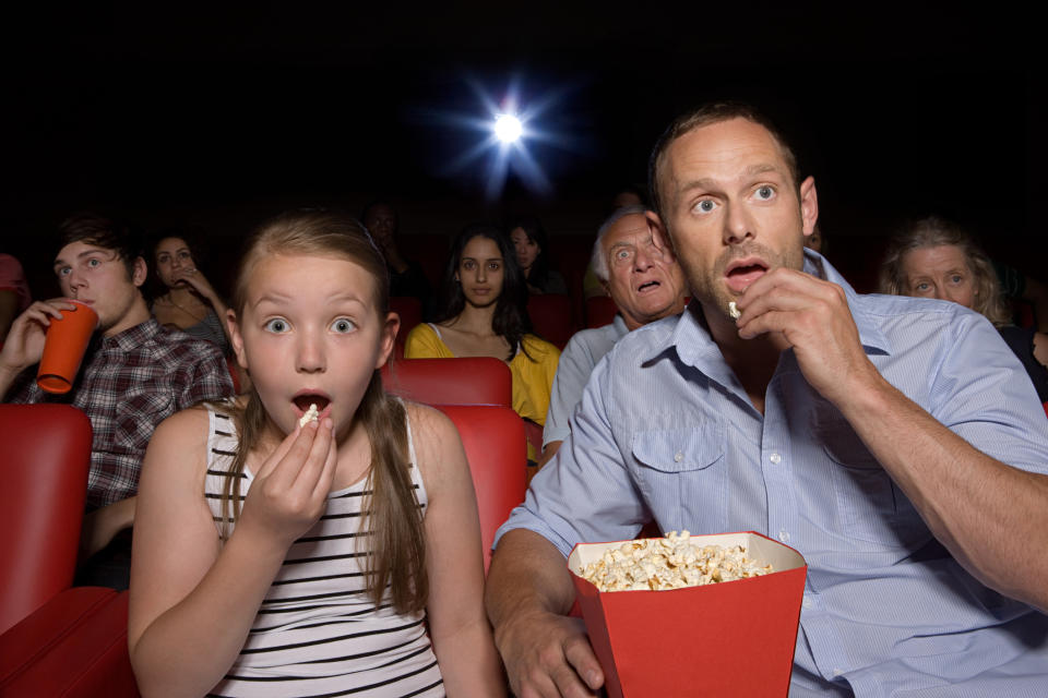 Father and daughter sharing a bucket of popcorn in a packed movie theater.