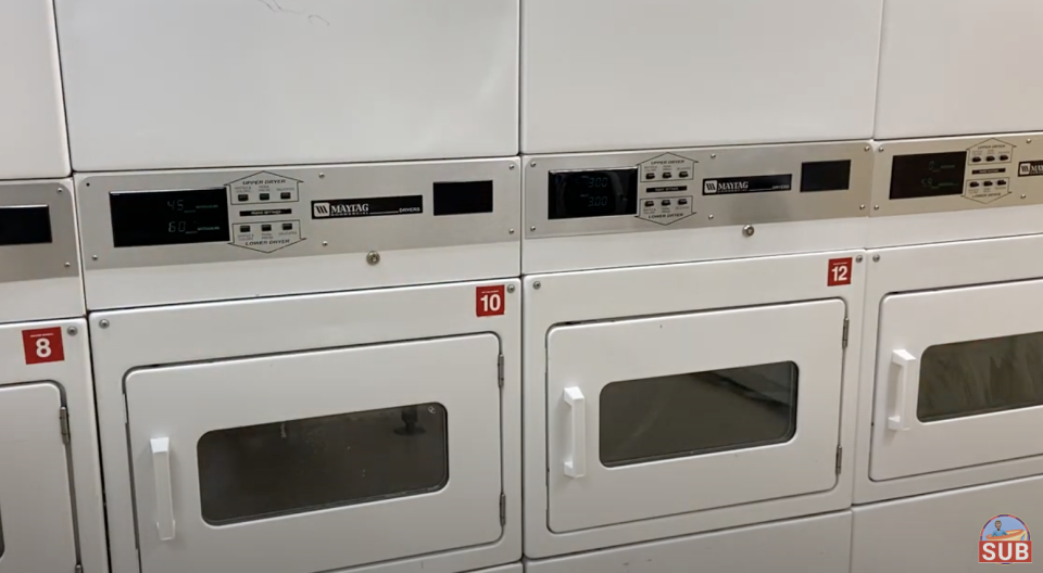 Four white commercial dryers in a laundromat are stacked two-high. Doors are closed. A small logo "SUB" is seen in the bottom right corner