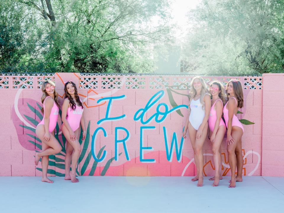 A group of women pose in front of a mural that says "I do crew."