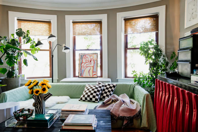 Living room with 3 windows with shades partly drawn, sofa with green cover and black and white checkered throw pillows, sunflowers on coffee table in black and white striped vase, large potted plants