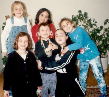 A group of smiling children, with a boy in the front wearing large earrings and many necklaces