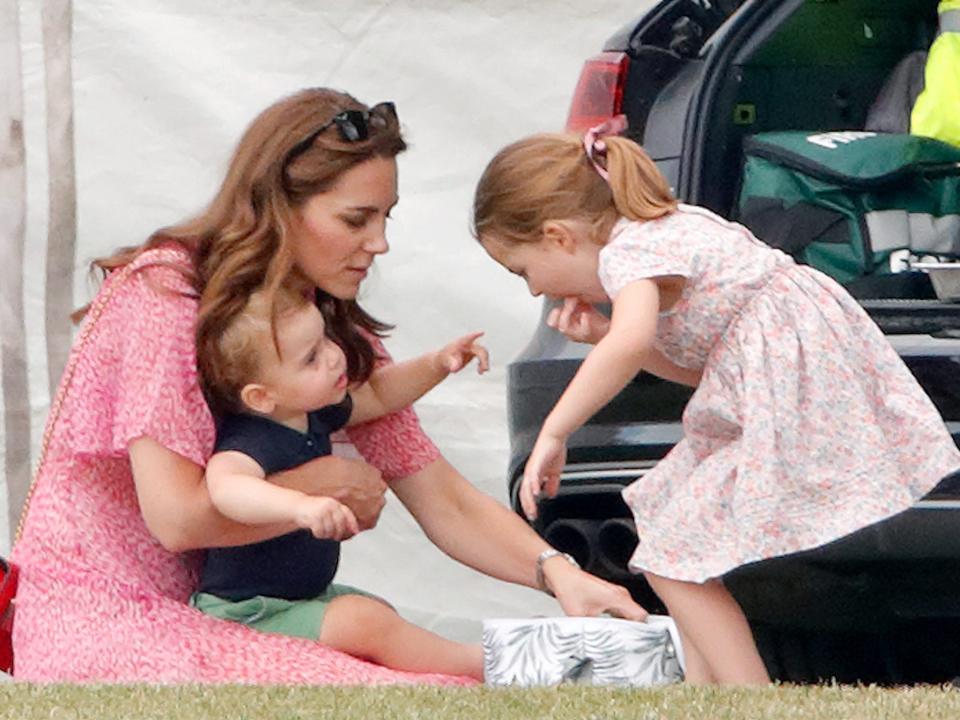 Kate Middleton sits on the ground with Prince Louis on her lap as Princess Charlotte leans down to talk to them with a minivan in the background.