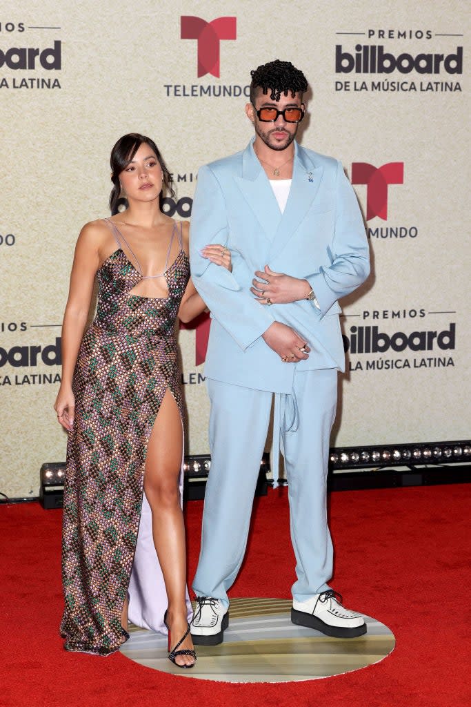 on a red carpet, Gabriela in a metallic gown with cut-outs, Bad Bunny in a pastel suit, both posing for cameras