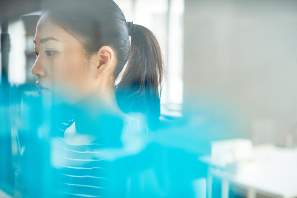 A woman with a ponytail is seen in profile through a glass panel, appearing thoughtful and focused. She is wearing a striped top