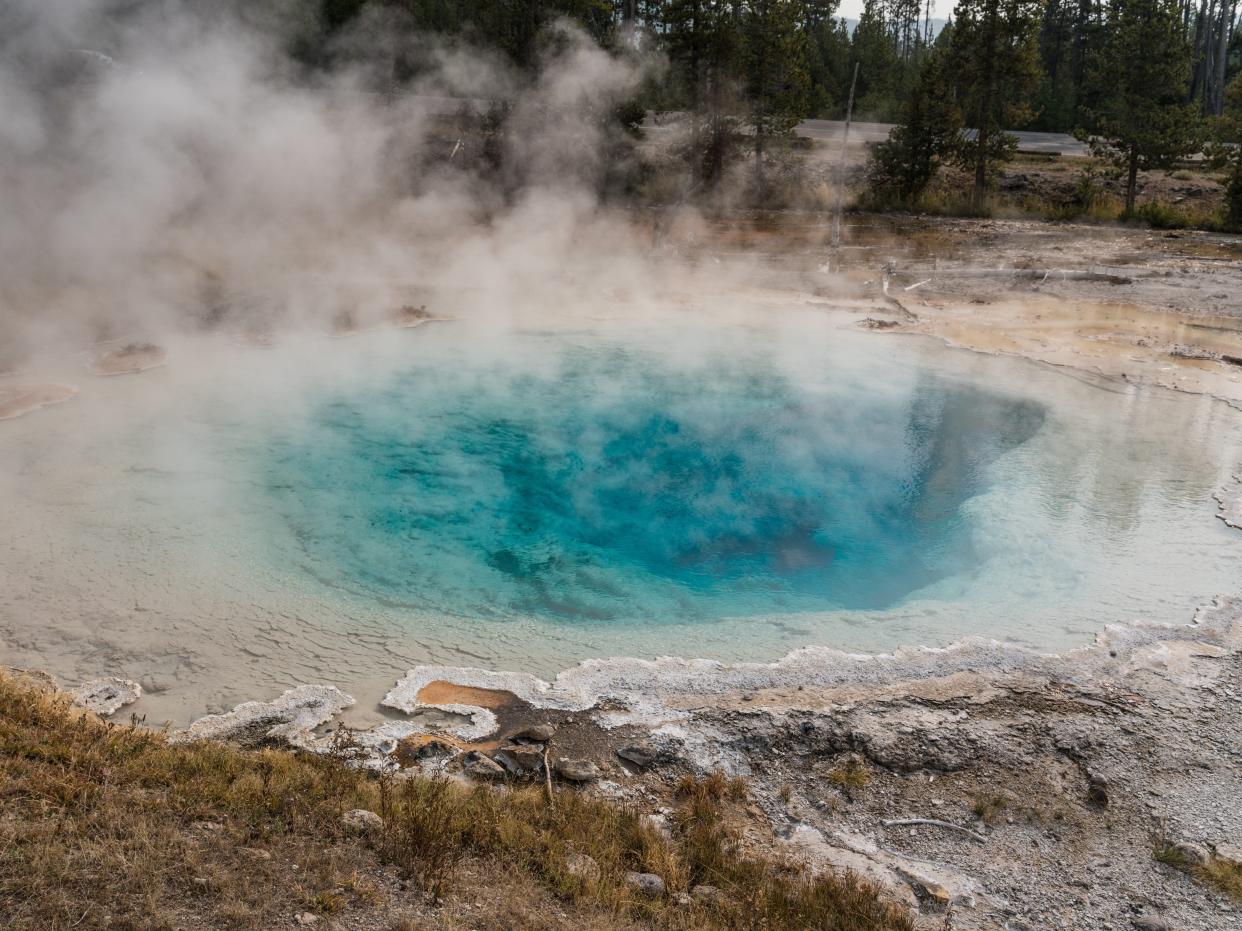 Steam rises off a large hot spring with cyan-colored water in the middle of a forested area.