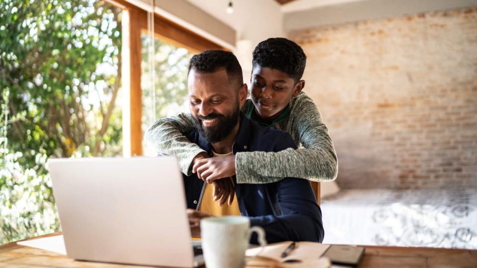 Smiling father uses laptop outside as son hugs him and looks over his shoulder at the screen