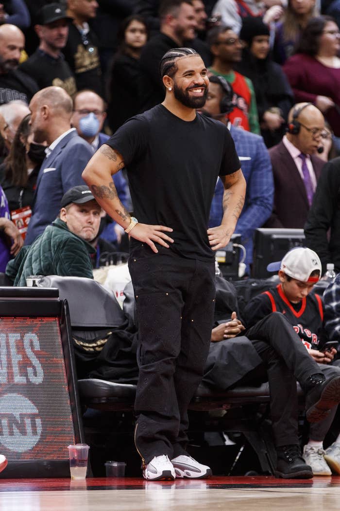 Drake standing courtside and smiling with his hands on his hips