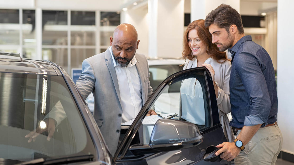 Mature african salesman showing all the car features to young couple.