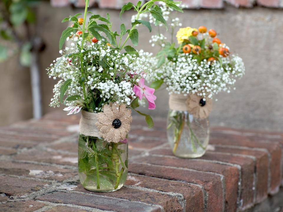 Two mason jars with burlap around them sit on a brick wall. Flowers sit in the jars.