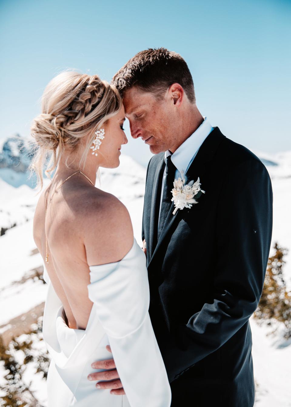 A bride and groom touch foreheads on top of a snowy mountain.