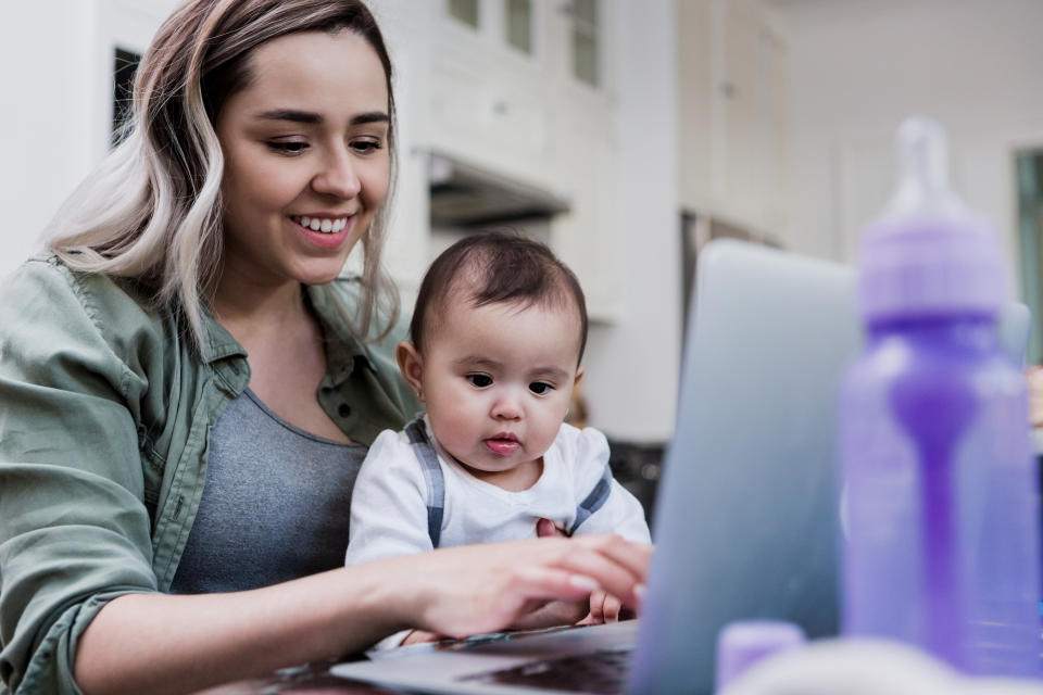 The mid adult mother uses her laptop to work from home as she holds her baby in her lap.