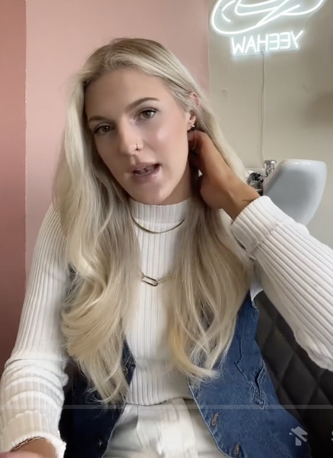 Woman with long blond hair in a ribbed top, touching earring, sitting in a salon chair