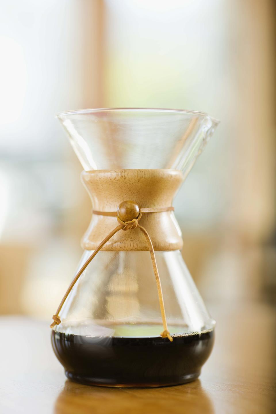 A glass Chemex coffee maker with a wooden collar and leather tie filled with brewed coffee sits on a table