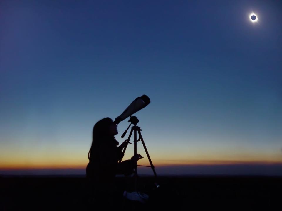 A person looks through a telescope at a total solar eclipse with sunset colors on the horizon behind her.