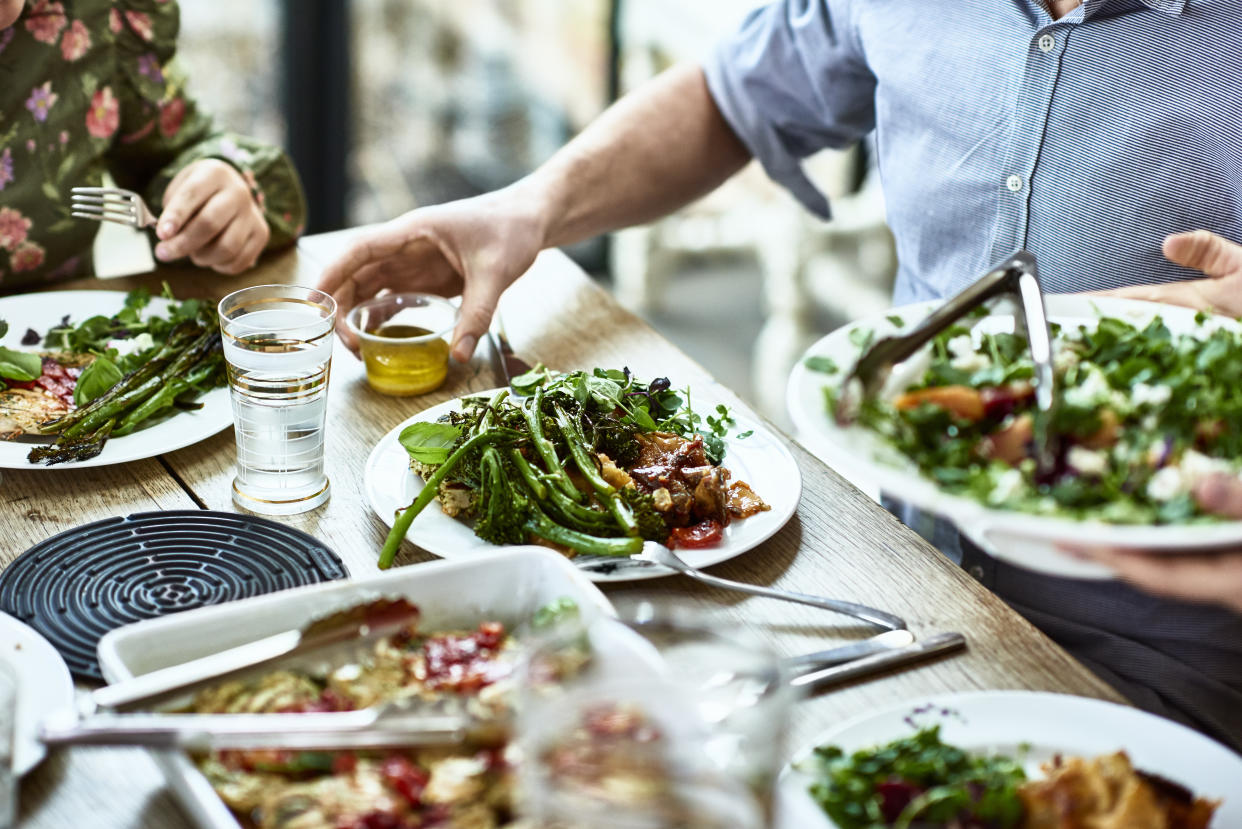 Close up shot of people sitting at dining room table with plates of vegetables and salad, person serving guests at dinner party, man about to pour dressing on his meal