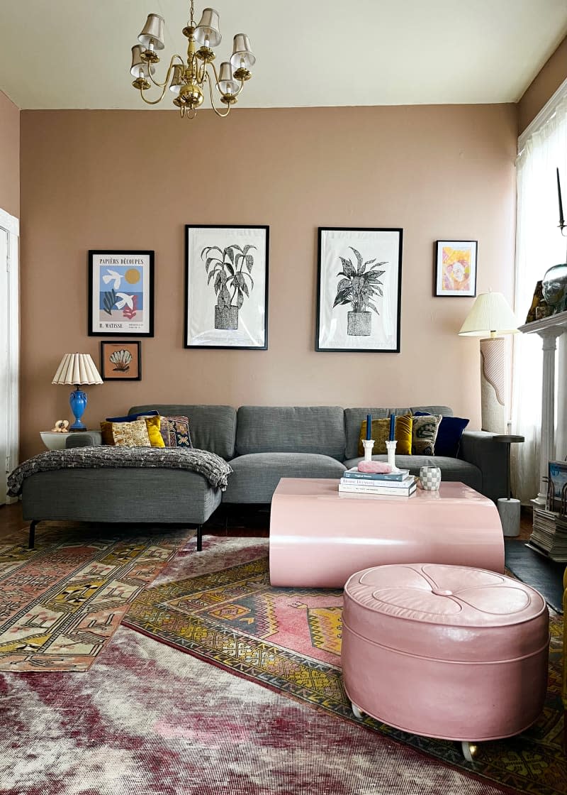 Gray couch and pink coffee table and ottoman sit atop multiple area rugs in living room with plant artwork hung on pink brown wall.