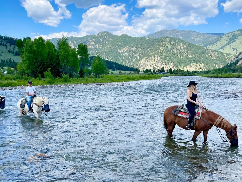 Photo of Melissa Persling from the side horseback riding, while crossing a wide river. Her horse is brown with a light blonde mane and black tail and leans down to drink from the river. Melissa has long blonde hair and wears a black boho-style hat, sunglasses, a black tank top, black pants, and boots. She holds the reins and sits on a Western-style saddle. Behind her, a few feet away, her ex-boyfriend Jim is riding a white horse with a white mane. Jim wears a blue baseball cap, white-shirt, and light blue denim jeans. In the background is a wide landscape featuring a bright green tree-line, a tree-strewn mountain range and bright blue sky with big white clouds.