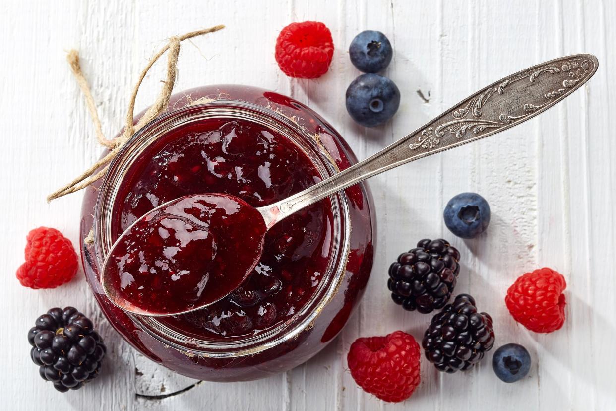 Top view of a spoon with wild berry jam over a glass jar of wild berry jam on a white wooden table with scattered berries