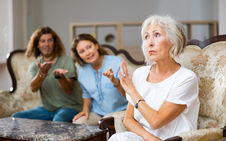 Elderly woman looking annoyed, raising her hand as if to stop the conversation, while a man and a woman in the background appear frustrated and pleading