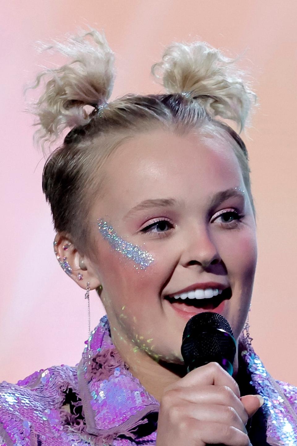 JoJo Siwa speaking into a microphone, wearing a sparkly purple jacket and hair in a bun with a bow