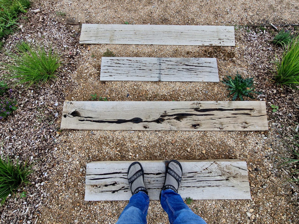 A person stands on a wooden pathway in a garden, wearing sandals and jeans; grassy plants surround the path