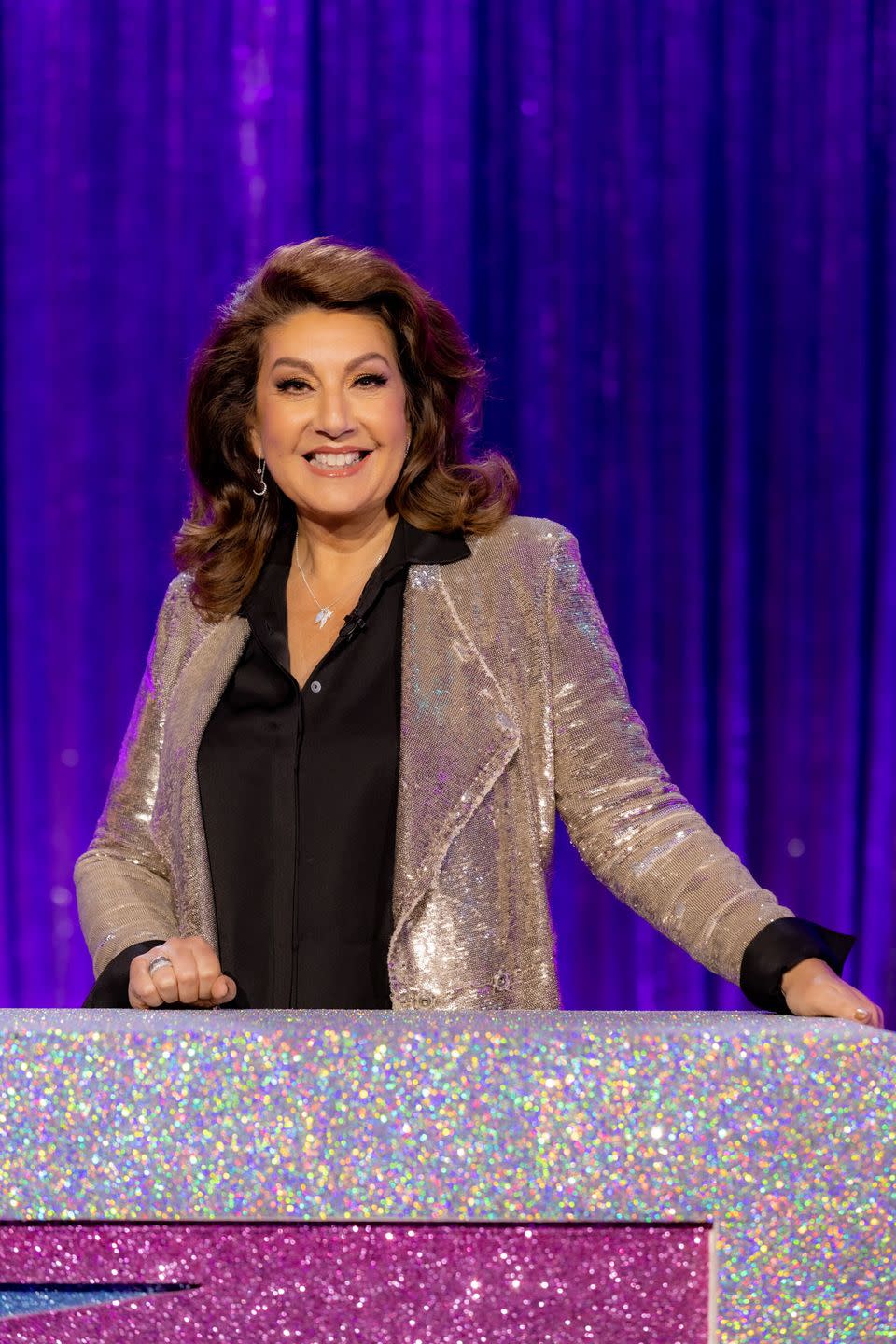 jane mcdonald in drag race vs the world season 2, an older woman stands behind a desk smiling at the camera, she has shoulder length curly brown hair and wears a black blouse with gold sequin jacket