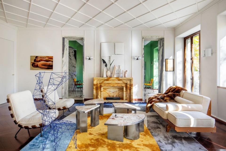 living room with white walls and ceiling with square panels, ocher colored marble fireplace, blue and ocher rug, white boucle chairs and sofa, blue wire chair, metal based cocktail tables with marble tops