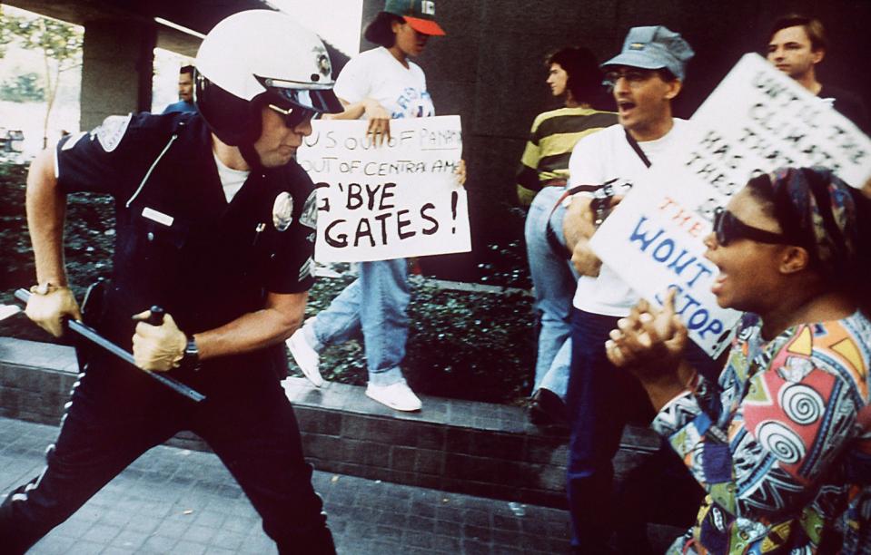 An LAPD officer grips his baton with two hands, preparing to strike a screaming woman