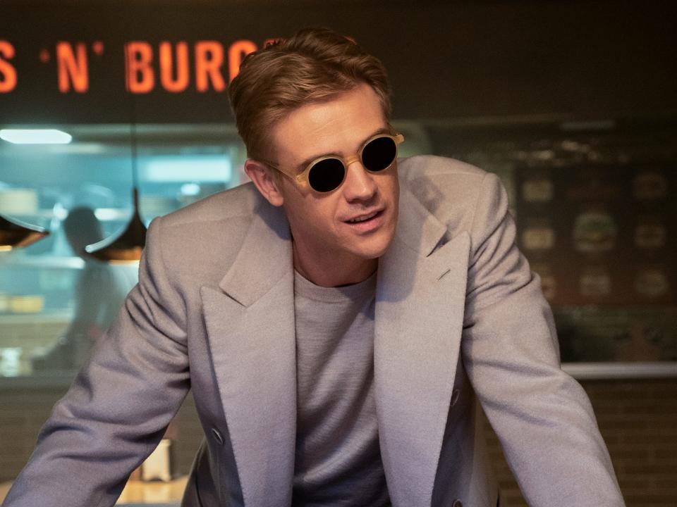 boyd holbrook as the corinthian in the sandman, leaning over a table in a diner. he has short blonde hair, and is wearing a grey blazer over a grey t-shirt, and circular sunglasses
