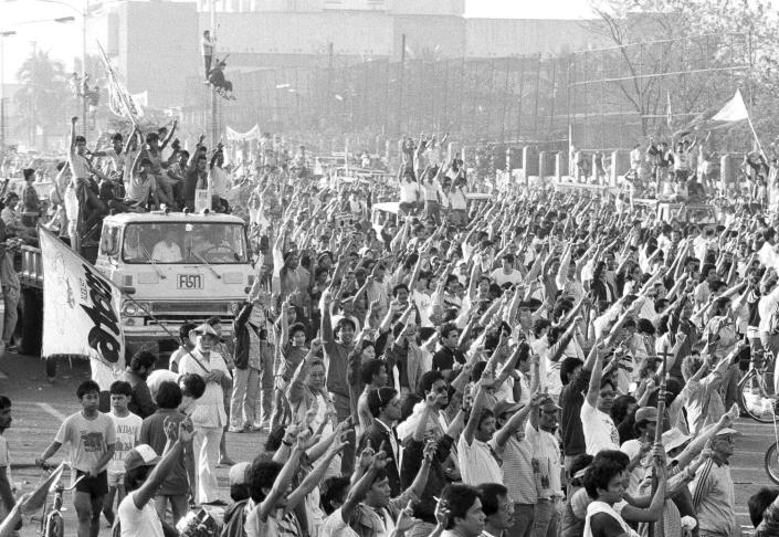 A black-and-white photo shows a vehicle carrying people with raised arms, next to a large crowd with outstretched arms