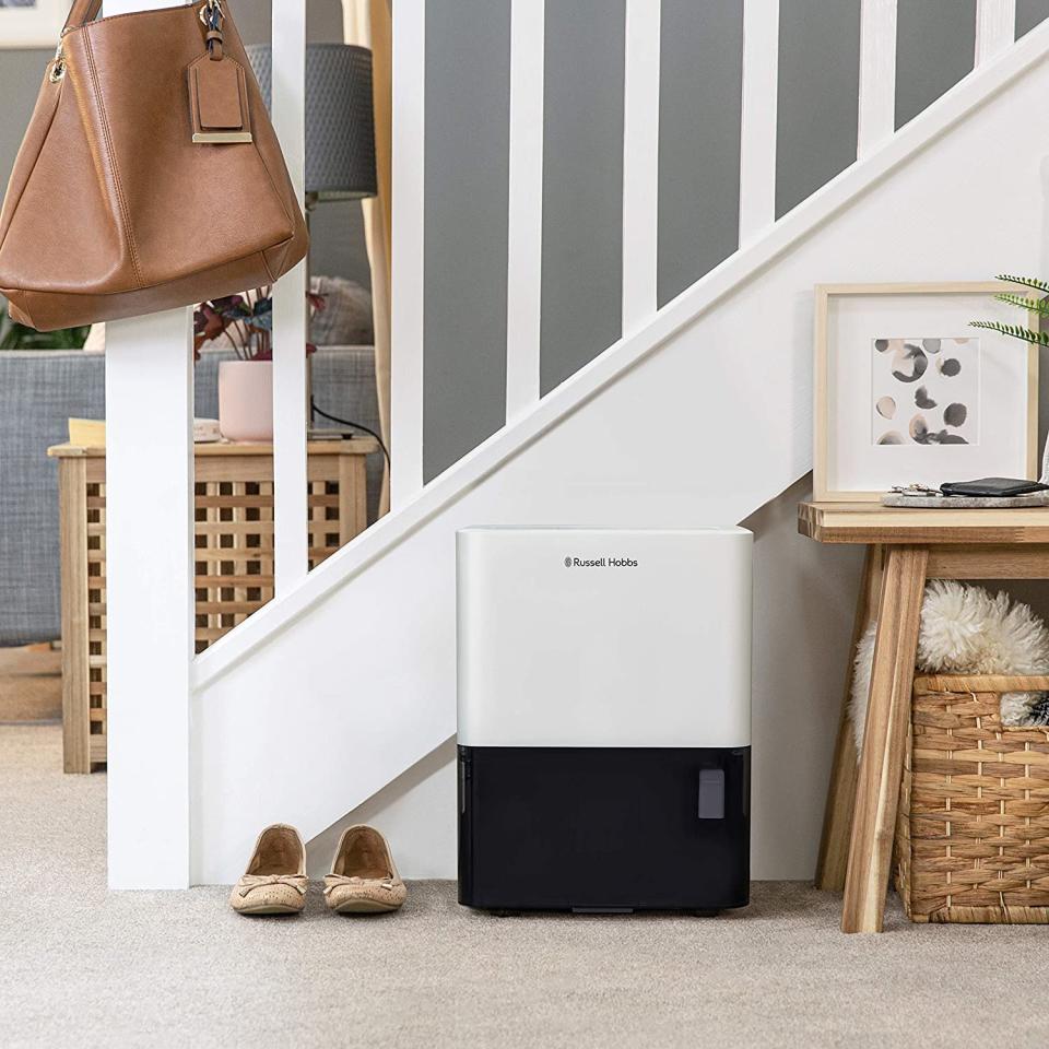 A household scene at the base of white stairs with a black and white dehumidifier sitting on beige carpet near shoes and sidetables. It is a black and white Russell Hobbs 2 Litre Dehumidifier, $271.62
